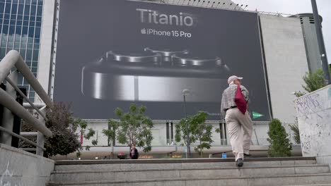 An-Einem-Einkaufszentrum-Hängt-Eine-Große-Kommerzielle-Plakatwerbung,-Die-Das-Apple-IPhone-15-Pro-Zum-Verkauf-Ankündigt,-Während-Ein-Käufer-Am-Rahmen-Vorbeigeht