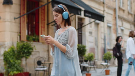 Mujer-Joven-Feliz-Con-Auriculares-Inalámbricos-Eligiendo,-Escuchando-Música-En-Un-Teléfono-Inteligente-Bailando-Al-Aire-Libre