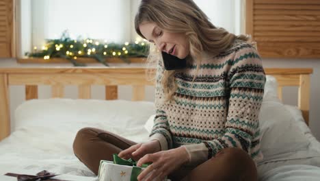 Caucasian-woman-wrapping-Christmas-gift-and-talking-on-phone-while-sitting-on-bed