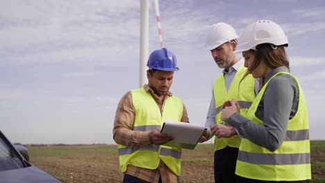 Three-caucasian-and-latin-engineers-standing-on-wind-turbine-field-and-discussing-over-plastic-models.
