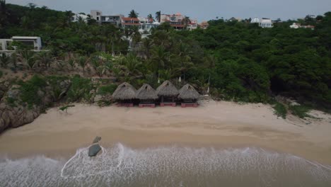 Aerial-view-of-some-beachfront-cabins-in-Huatulco-at-the-end-of-the-day-while-the-waves-are-crashing-on-shore