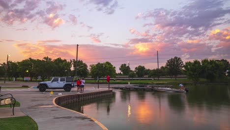 Time-lapse-of-man-fishing-from-pier-with-net-and-pole-as-truck-drops-boat-off-at-ramp,-sunrise-sky-reflected-in-water