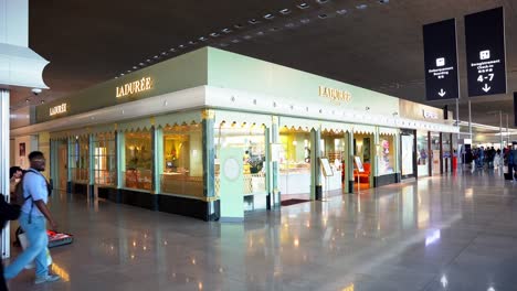 Ladurée-luxury-pastry-shop-at-Paris-airport,-giant-modern-French-design,-tourists-walking-along-the-front-of-the-shop