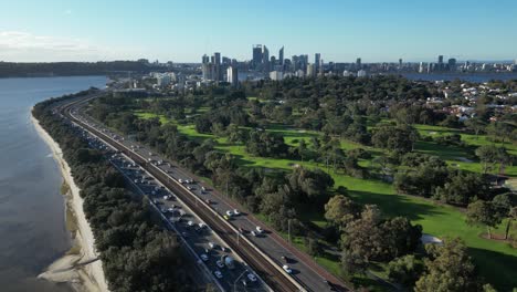 Drone-backwards-shot-of-busy-traffic-on-coastal-road-next-to-golf-course-and-Perth-City-in-Background