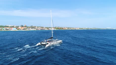Catamaran-rocking-over-blue-open-ocean-waves-at-midday,-drone-follows-from-behind