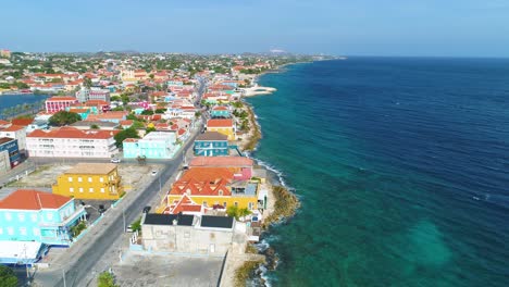 Vibrant-coastal-homes-of-Willemstad-Curacao-near-Punda-District,-trucking-pan-out-to-sea-as-waves-crash-on-rocky-coast