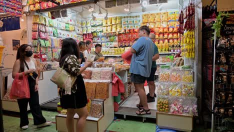 Two-ladies-checking-out-a-food-shop-selling-snacks-and-colorful-goodies-at-a-market-while-other-customers-wait-for-their-purchases-in-Chatuchak,-Bangkok,-Thailand