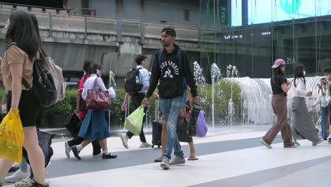 Local-and-foreign-tourists-walking-beside-a-water-fountain-at-the-walkway-in-between-Siam-Paragon-Mall-and-the-BTS-skytrain-station,-in-Bangkok,-Thailand