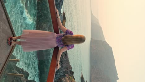 Beautiful-woman-stands-at-glass-corner-overlook-sea-cliff-vista,-waves-crash-in-slow-motion,-vertical