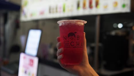 A-hand-is-holding-up-a-plastic-cup-of-local-Thai-bubble-tea-in-front-of-a-street-vendor-stand-in-Bangkok