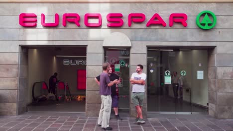 Three-men-stand-in-front-of-a-supermarket-and-talk-to-each-other-while-people-walk-past