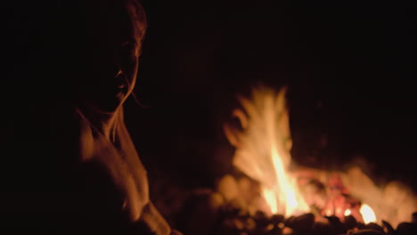 Campfire-light-illuminates-shirtless-man-on-dark-evening,-fire-in-background-out-of-focus
