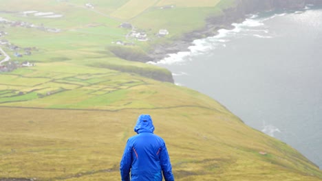 Back-view-of-man-in-wet-blue-jacket-balancing-on-one-foot-in-Villingardalsfjall,-Faroe-Islands