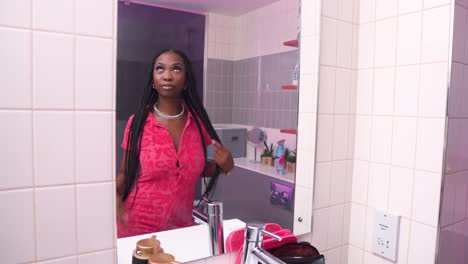 Afrikanische-Model-Schauspielerin-Trägt-Ein-Rosafarbenes-Kleid-Mit-Knöpfen-Und-Langen-Zöpfen-Im-Badezimmer-Im-Badezimmer-Vor-Dem-Spiegel-Und-überprüft-Sich-Selbst.-Sie-überlegt,-Was-Sie-Sonst-Noch-In-Ihre-Make-up-Tasche-Stecken-Soll.-Modernes-Badezimmer-Im-Kino