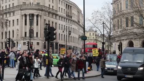 Pedestrians-crossing-street-on-crosswalk-in-London-city-with-red-bus-and-people-protesting-at-Trafalgar-Square