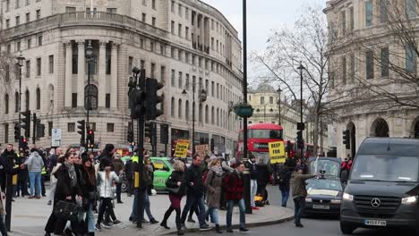 Los-Peatones-Cruzan-La-Calle-En-El-Paso-De-Peatones-En-La-Ciudad-De-Londres-Con-El-Autobús-Rojo-Y-La-Gente-Protestando-En-Trafalgar-Square.