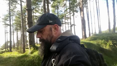 A-bearded-man-in-a-baseball-cap-strolls-leisurely-along-a-sunny-trail-in-the-woodland-forest-of-Newborough,-Isle-of-Anglesey,-Wales