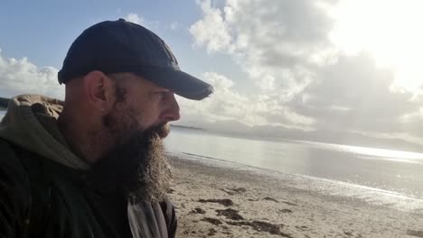 A-thinking-man-with-a-beard-and-cap-is-walking-along-Newborough-Beach-on-a-beautiful-sunny-day,-with-sunbeams-reflecting-like-a-mirror-in-the-water,-Isle-of-Anglesey,-Wales