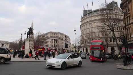 El-Tráfico-De-La-Ciudad-De-Londres-En-La-Rotonda-De-Trafalgar-Con-Taxis-Negros-Y-Autobuses-Rojos-De-Dos-Pisos-Pasando-Por-Personas-Que-Protestan