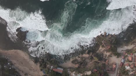 Small-village-on-coastline-with-powerful-ocean-waves,-aerial-top-down-view