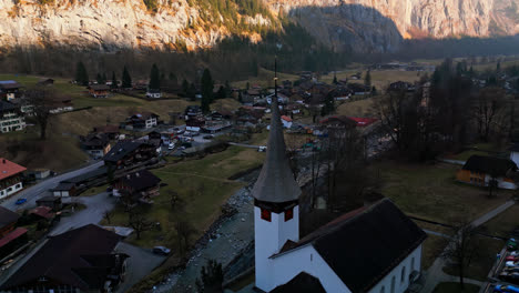 Drone-pulls-back-from-chapel-spire-to-reveal-serene-town-of-Lauterbrunnen,-Switzerland-deep-in-the-canyon