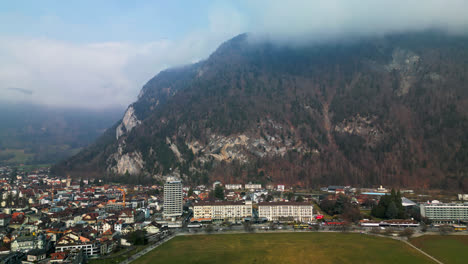 Drone-ascends-showcasing-sprawling-town-of-Interlaken-Switzerland-at-the-base-of-a-forested-mountain-in-the-clouds