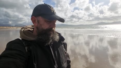 Bearded-male-wearing-baseball-cap-slow-motion-walking-windy-beach-reflecting-clouds