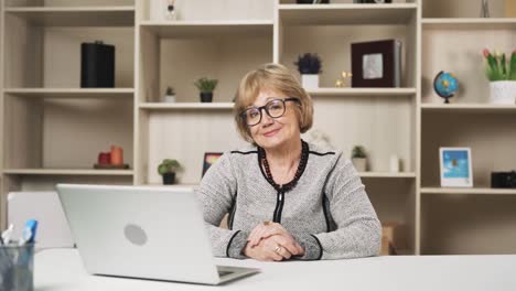 elderly-woman-puts-on-glasses-and,-smiling,-looks-into-the-camera-while-sitting-at-the-table-with-a-laptop