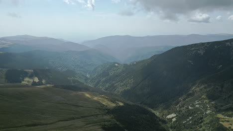Captivating-aerial-view-of-the-serene-Transalpina-region-in-Romania,-showcasing-vast-forested-valleys,-shadowed-mountain-ranges,-and-untouched-open-meadows-under-a-soft-clouded-sky