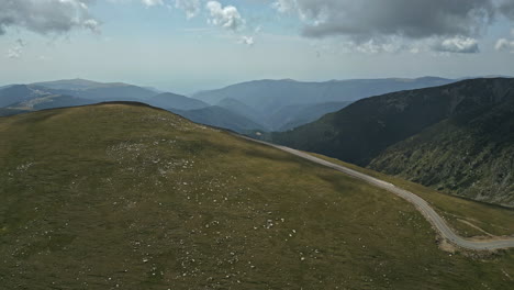 Majestätisches-Panorama-Der-Transalpina-Rumäniens,-Das-Weite-Grüne-Hochebenen-Und-Eine-Einsame-Straße-Hervorhebt,-Die-Sich-Durch-Sie-Schlängelt,-Mit-Fernen-Bergschichten-Unter-Einem-Gesprenkelten-Himmel