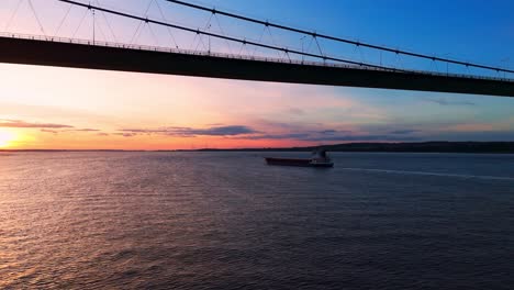 Embark-on-a-serene-journey-as-a-barge-boat-leisurely-cruises-beneath-the-Humber-Bridge-during-sunset,-enveloped-in-the-golden-hues-of-the-setting-sun,-with-the-river-awash-in-shimmering-light