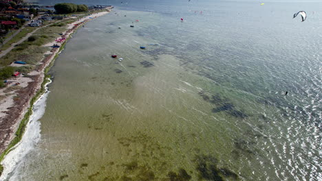 Aerial-view-of-Kuźnica-beach,-showcasing-kitesurfers-gliding-over-glistening-waters,-adjacent-to-a-serene-coastal-town