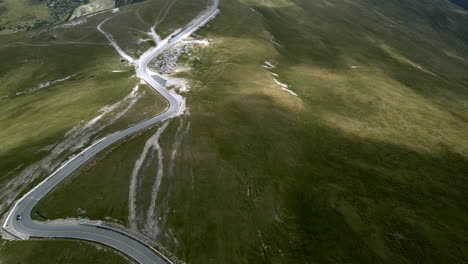 Aerial-shot-of-the-winding-Transalpina-road-in-Romania,-gracefully-cutting-through-lush-green-landscapes-with-vehicles-in-motion,-embodying-scenic-drives