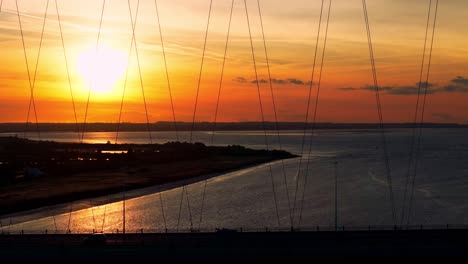 A-tranquil-scene-unfolds-as-a-barge-boat-glides-serenely-under-the-Humber-Bridge-during-sunset,-casting-a-warm,-golden-hue-upon-the-shimmering-river,-captured-in-breathtaking-aerial-footage
