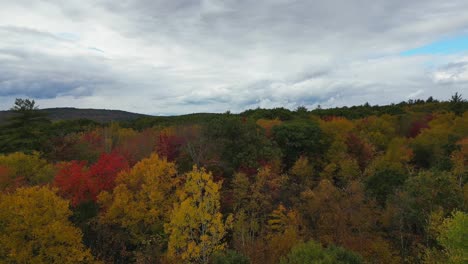 Aerial-over-Massachusetts'-autumn-forests,-vibrant-orange-red-trees-contrast-with-the-rain-filled-clouds-and-the-touch-of-blue-sky