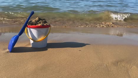 Full-toy-bucket-with-blue-scoop-on-sandy-beach-with-waves-breaking-on-shore