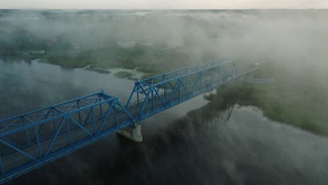 Aerial-establishing-view-of-the-steel-bridge-over-Lielupe-river-on-a-sunny-summer-morning,-fog-rising-over-the-river,-cars-driving,-wide-ascending-drone-shot,-tilt-down