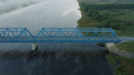 Aerial-establishing-view-of-the-steel-bridge-over-Lielupe-river-on-a-sunny-summer-morning,-fog-rising-over-the-river,-cars-driving,-wide-drone-dolly-shot-moving-left