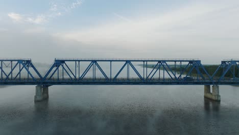 Aerial-establishing-view-of-the-steel-bridge-over-Lielupe-river-on-a-sunny-summer-morning,-fog-rising-over-the-river,-cars-driving,-wide-drone-dolly-shot-moving-left-low