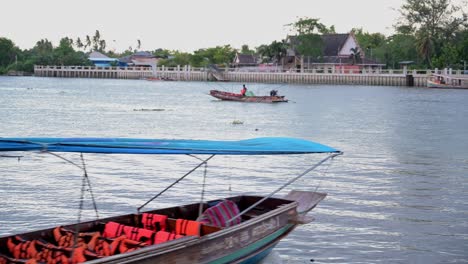 Ein-Bootsmann,-Der-Mit-Seinem-Langen-Ausflugsboot-Den-Fluss-Befährt,-Während-Ein-Anderer-Mann-Mit-Seinem-Boot-Aus-Der-Ferne-Ganz-In-Der-Nähe-Des-Schwimmenden-Marktes-Amphawa-In-Samut-Songkram,-Thailand,-Zu-Sehen-Ist