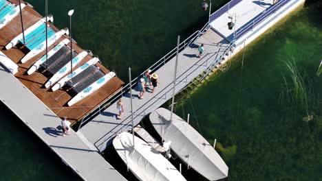 Gente-Ocupada-Caminando-Por-Un-Paseo-Marítimo-Flotante-Junto-A-Aguas-Cristalinas-Y-Veleros