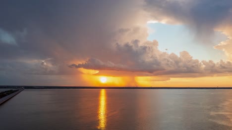 Sun-sets-and-reflections-golden-yellow-ray-across-lake,-storm-clouds-billowing-and-growing