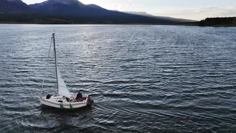 sailboat-moving-in-the-wind-on-dillon-dam-reservoir-at-sunset-in-the-shade-with-snow-covered-mountains-in-the-background-AERIAL-ORBIT