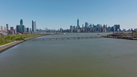 Aerial-backwards-shot-of-river-with-bridge-and-park-in-New-York-City-at-sunny-day---Skyline-of-Big-Apple-in-background