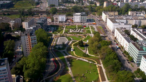 Aerial-shot-of-an-urban-area-showcasing-modern-buildings,-a-landscaped-park-with-winding-pathways,-and-busy-streets-with-moving-vehicles
