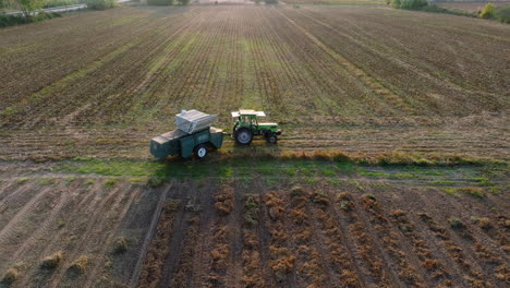 Aerial:-Side-follow-of-a-tractor-with-trailer-in-an-empty-field