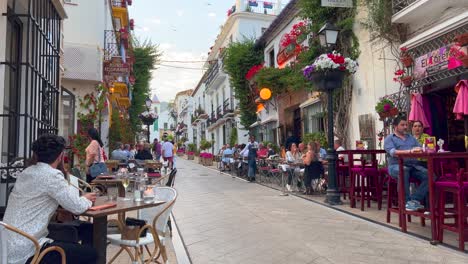 People-sitting-and-having-food-outside-in-restaurants-in-Marbella-old-town-Spain,-busy-romantic-street-during-summer,-tourists-walking,-4K-static-shot