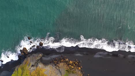 Top-down-aerial-view-of-the-black-sand-beach-in-southern-Iceland,-rocky-coastline-with-waves-splashing-the-black-sand-beach