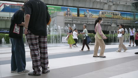 People-are-busy-walking-outside-of-Siam-Paragon-in-Bangkok-with-the-BTS-train-station-with-trains-leaving-and-stopping-for-passengers,-in-Thailand
