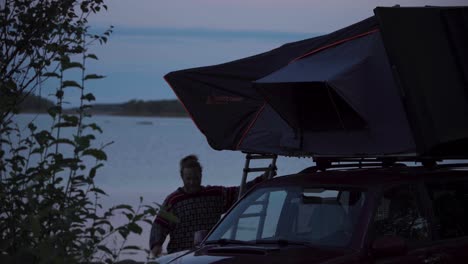 Un-Hombre-Baja-Por-La-Carpa-Del-Techo-Del-Auto-Cerca-De-La-Orilla-Del-Lago-Al-Atardecer